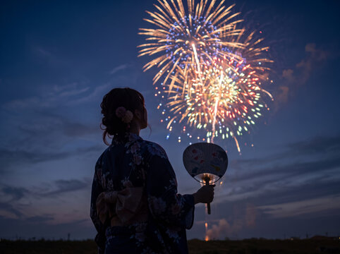 夏空に咲く大輪の打ち上げ花火とうちわを持つ浴衣姿の女性