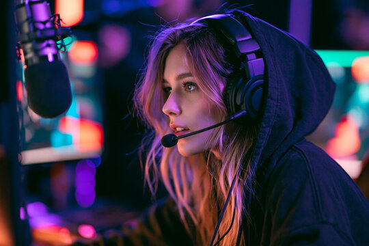 A focused young woman wearing a headset and hoodie streams content in a dimly lit studio illuminated by vibrant neon pink and blue ambient lighting