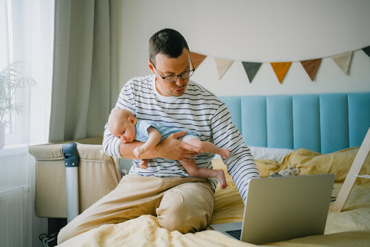 Father balancing a newborn baby while working on a laptop from bed, representing the challenges and realities of remote work, paternity leave, and modern parenting responsibilities at home