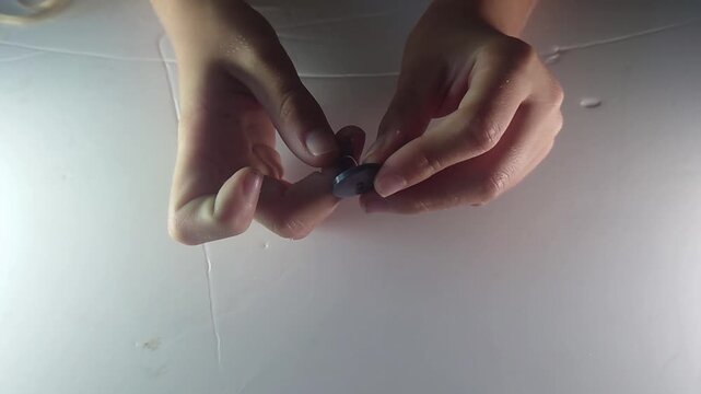 A top-view close-up shot shows human hands carefully threading a thin string through the holes of a dark button to construct a traditional vintage whirlygig spinner toy on a bright white surface.
