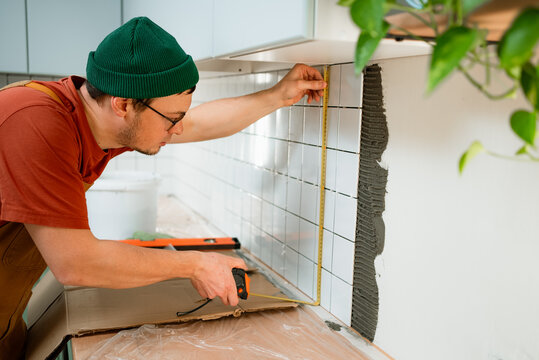 Man wearing a green beanie measuring and affixing white ceramic tiles to a kitchen wall, meticulously working on a home improvement backsplash renovation project with precision
