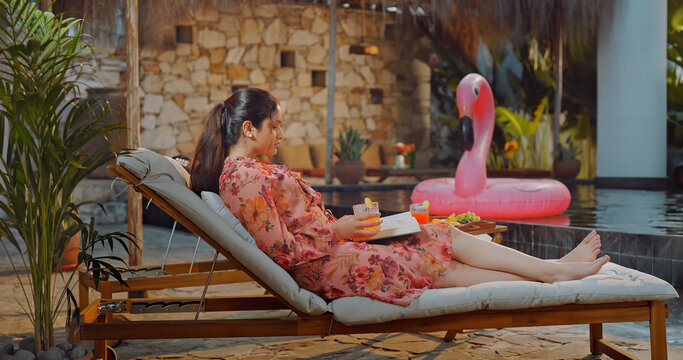 Indian young adult relax woman sitting on sun lounger chair read new book enjoy picnic trip day happy girl hold casual novel do self care spend time have fun joy villa pool edge place