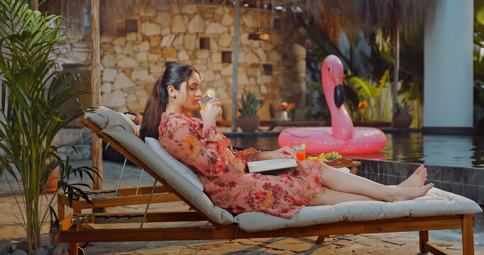 Indian young adult relax woman sitting on sun lounger chair read new novel book enjoy picnic trip day happy girl hold glass drink casual juice do self care spend time have fun joy pool edge place