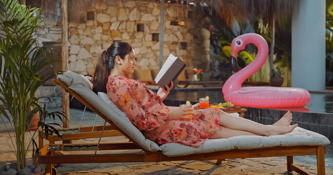 Indian young adult relax woman sitting on sun lounger chair read new book enjoy picnic trip day happy girl hold casual novel do self care spend time have fun joy villa pool edge place