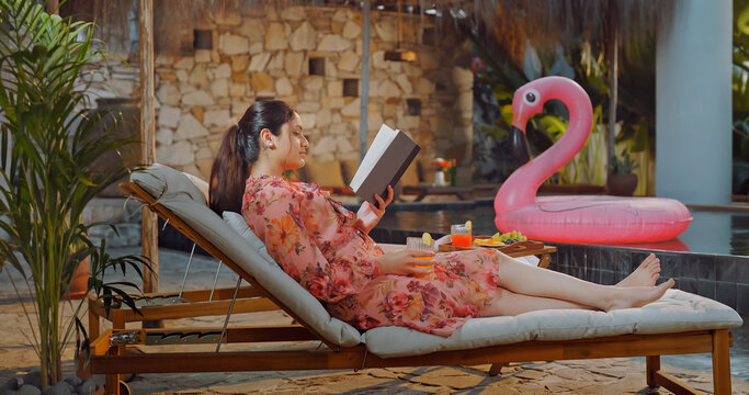 Indian young adult relax woman sitting on sun lounger chair read new book enjoy picnic trip day happy girl hold casual novel do self care spend time have fun joy villa pool edge place