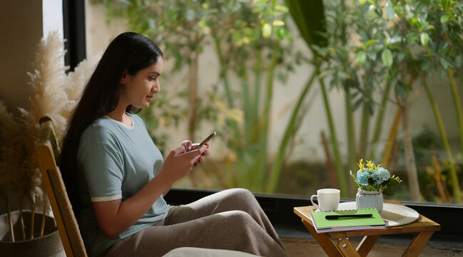 Indian young adult gen z relax female girl sitting on chair use mobile do virtual chat take job break room happy resting woman hold phone scrolling social media video enjoy day spend time home place
