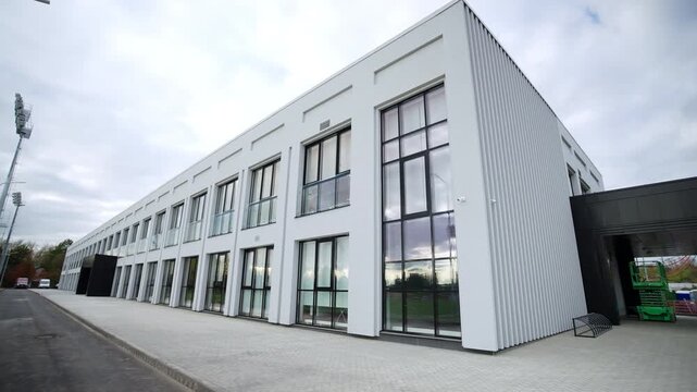 Modern building facade features white vertical corrugated panels. Large glass windows reflect green trees bright sky on overcast day