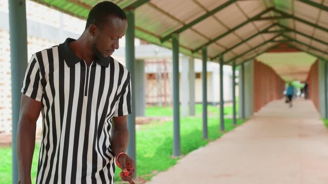 Black commuter closeup in covered walkway striped polo looking down at hand accessory shallow depth vanishing point perspective muted tones reflective mood campus background late morning light