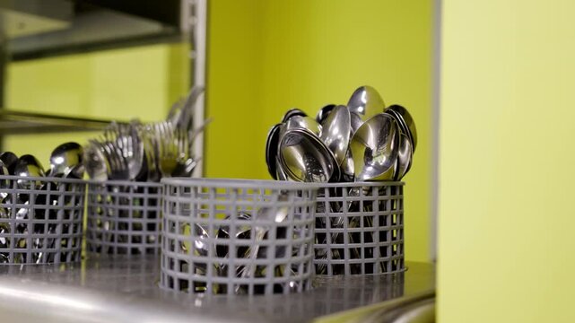 Clean metal spoons and forks sit in plastic baskets. Polished stainless steel counter reflects bright kitchen lighting. Kitchen utensils