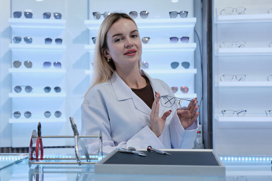Portrait of White female expert optometrist in medical coat holding eyeglasses, lens guidance, improved eyesight for individual lifestyles, professional care service at store display in optical shop.