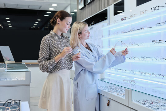 White female optometrist helps and guides customers to choose eyeglasses for improved eyesight with individual lifestyles, consults on decisions, and options choices at store display in optical shop.