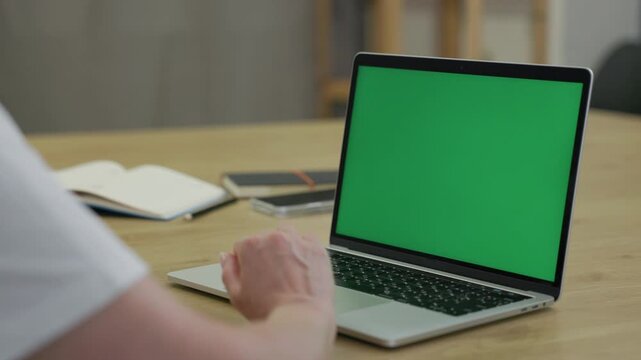 Woman Working On Laptop Computer with Green Mock Up Screen At Home Office Space. Female Freelancer Browsing Green Laptop Screen Writing Information in Notebook