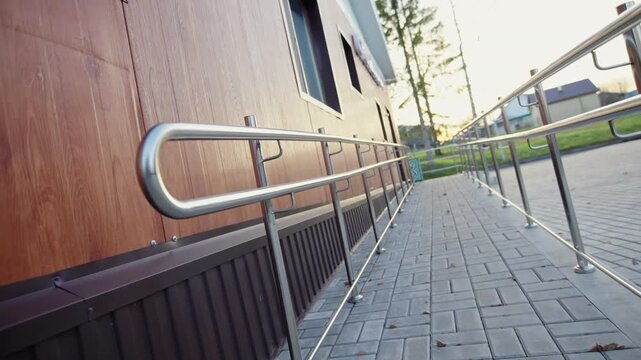 Metal shiny handrails of outdoor ramp stand on paved pedestrian path.Contemporary building with wooden siding sits in close background