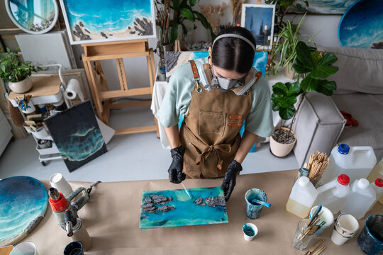 Woman crafter in apron and protective respirator creating epoxy resin art at worktable in workshop, female artisan in home studio uses tools and instruments, spreading liquid resin over canvas surface