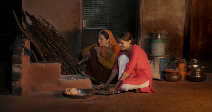 Indian chef mom teen female girl do work talk speak use clay mud stove indoor rural area young adult woman lady cook casual food on open fire flam make fresh roti chapati at dark room home desi life