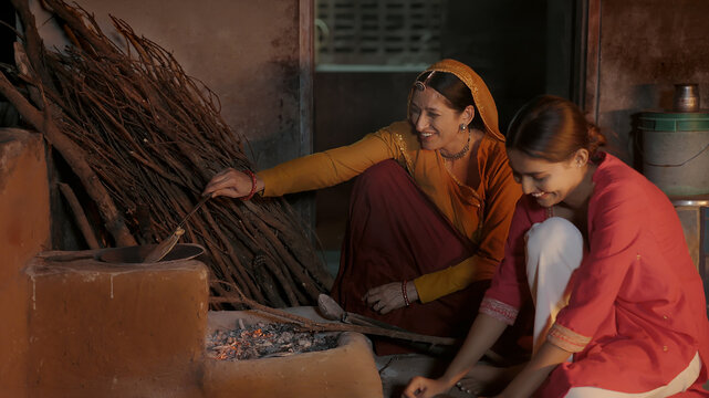 Indian chef mom teen female girl do work talk speak use clay mud stove indoor rural area young adult woman lady cook casual food on open fire flam make fresh roti chapati at dark room home desi life