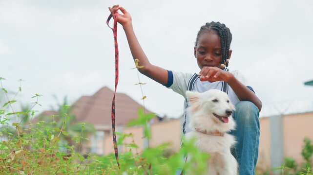 Black child sitting with white dog outdoors by suburban wall, girl holds leash and gazes thoughtfully into distance, calm moment of companionship, soft grass foreground and muted cloudy sky, intimate