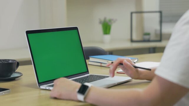 Woman Working On Laptop Computer with Green Mock Up Screen At Home Office Space. Female Freelancer Browsing Green Laptop Screen Writing Information in Notebook
