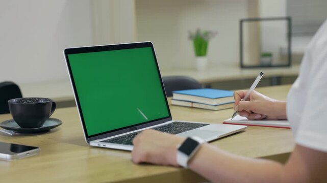 Woman Working On Laptop Computer with Green Mock Up Screen At Home Office Space. Female Freelancer Browsing Green Laptop Screen Writing Information in Notebook