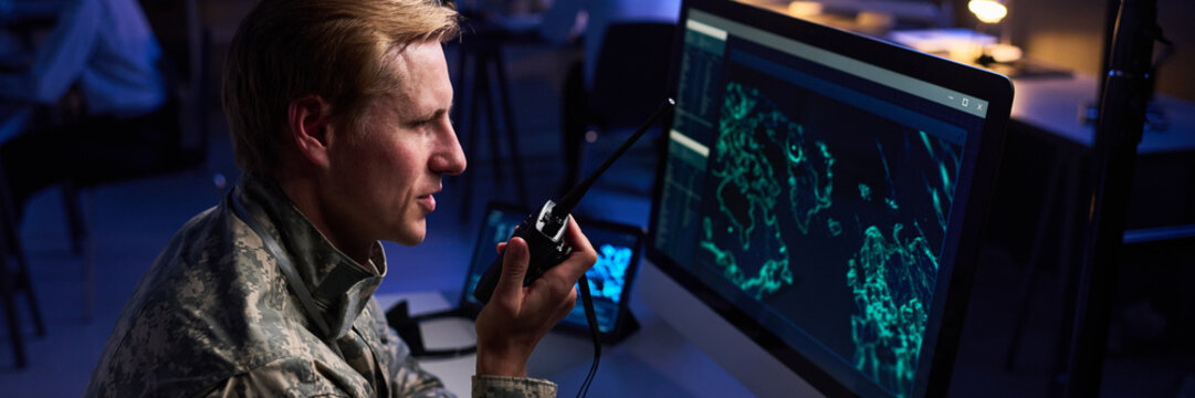 Caucasian young adult military officer speaking into walkie talkie while looking at computer screen with digital map. Image suitable for defense technology and security services marketing