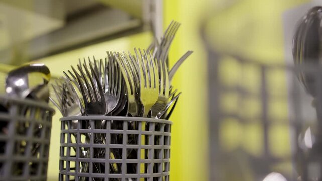 Polished metal cutlery organized on self-service cafeteria counter. Yellow background wall contrasts with plastic kitchenware holders