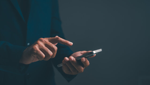 A person in a dark suit using a smartphone against a dark background, focusing on hand interaction with the device.