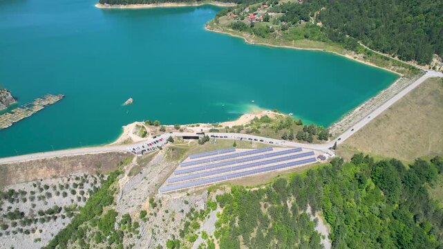 An aerial drone crosses Zaovine Lake in Serbia, revealing long rows of solar panels beside the dam road, turquoise water, wooded shoreline and a small mountain village in daylight.