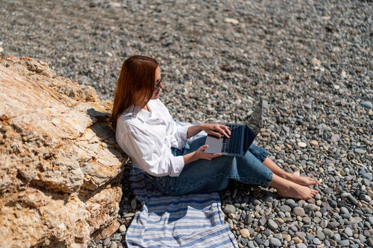 Remote work lifestyle featuring a woman using a laptop computer while sitting on a sunny pebble beach shore