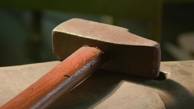 Blacksmith hammer head anvil macro detail illuminated, handheld shot. Extreme close up of metalworking tools inside workshop with warm lighting.