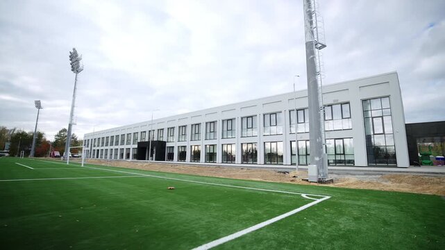 Tall floodlight masts rise high up against cloudy sky. Modern facility facade features multiple panoramic glass windows black doors