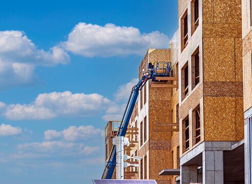 Multi-story wood frame building under construction with a blue boom lift reaching the upper floors against a bright blue sky with clouds.