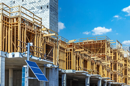A construction site features concrete foundation with extensive multi-story wooden framing for a large residential complex under a blue sky