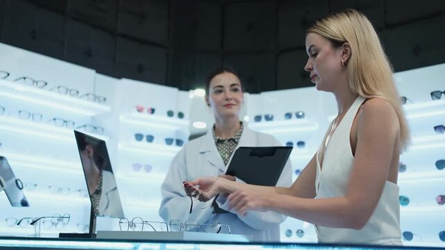 White female customer chooses eyeglasses with optometrist's help and guidance, improved eyesight for individual lifestyles, consults on decisions, and options choices at store display in optical shop.