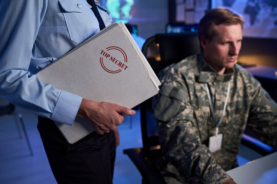 Female officer holding top secret folder near Caucasian military man working on computer in control room, representing national security and intelligence operations, medium shot