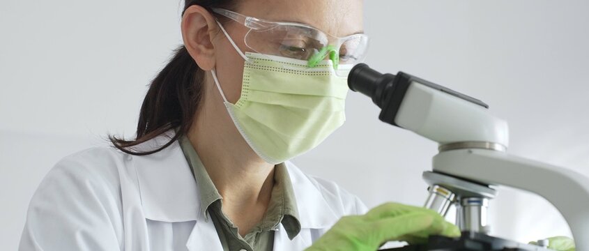 Female scientist examining specimen, placing slide under microscope while wearing green protective gear in sterile laboratory workspace. Medicine, healthcare and science concept