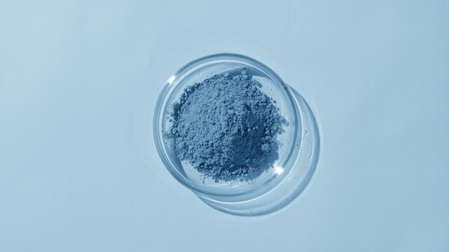 Top perspective capture of a pile of blue powder in a glass petri dish on a blue background, highlighted by rotating light casting shifting shadows | Woad powder cosmetic ingredient concept