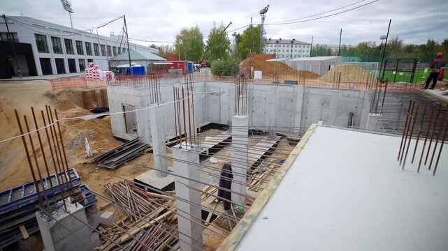 Formwork panels, wooden boards lie scattered on sandy ground. Distant white building trees border active urban infrastructure project site