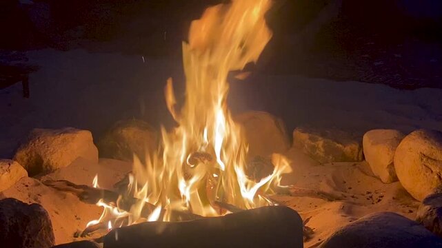 A vibrant campfire with bright orange and yellow flames burning in a circular stone fire pit set in the sand at night, creating a warm and cozy atmosphere.