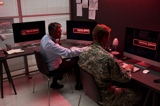 Middle aged Indian security guard and Caucasian soldier working on computers with critical error screens, managing cyber security breach. Useful for IT defense and crisis management ads