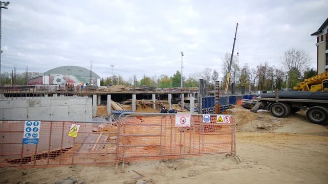 Active construction site features temporary orange safety fences. Multiple workers in helmets walk among concrete foundations columns