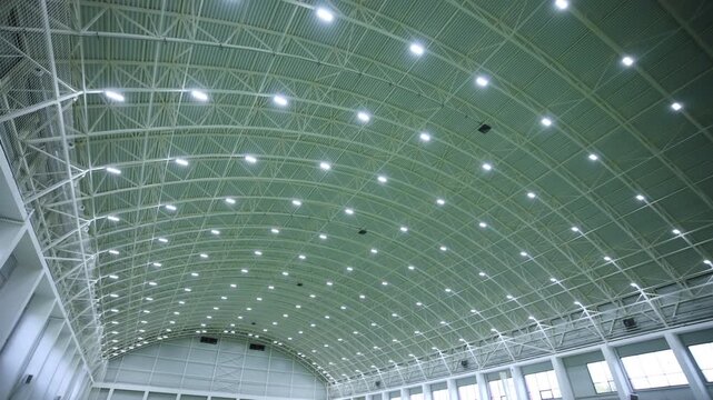 Massive arched ceiling structure decorates space inside modern sports arena. Dozens of bright lamps illuminate indoor sports field