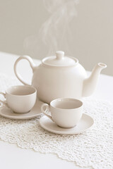 Fototapeta premium White ceramic teapot and two teacups rest on delicate lace as soft steam rises in a serene minimalist setting.