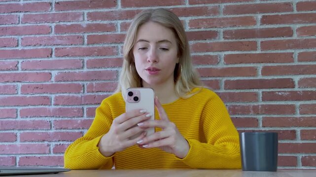 Caucasian blonde woman sitting at a desk with a brick wall background, actively using her smartphone for texting, browsing social media, and staying connected with friends online