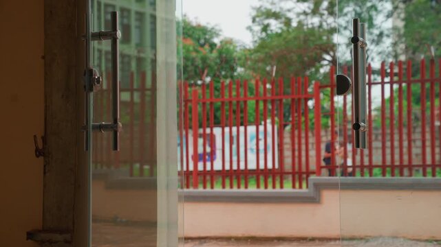 Open glass door framing red fence and quiet street, interior threshold with metal handle and latch in foreground, exterior yard and trees beyond fence, muted color palette and soft light, calm waiting