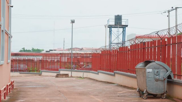 Red metal fence and courtyard outside family home, concrete walkway with dumpster, water tower and lamp posts in background, overcast sky, muted colors, practical urban protection suggesting safe