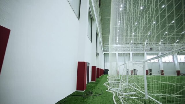 Arena wall shows red protective pads on columns and football goal netting. Modern soccer goal with net stands on green artificial turf
