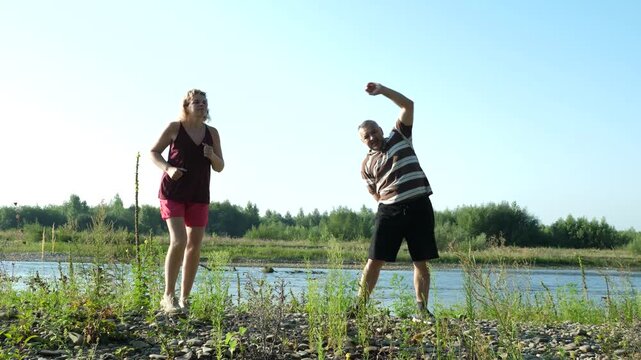 Two older adults exercise outdoors as a man performs a side bend while a woman does warm-up movements beside the river.