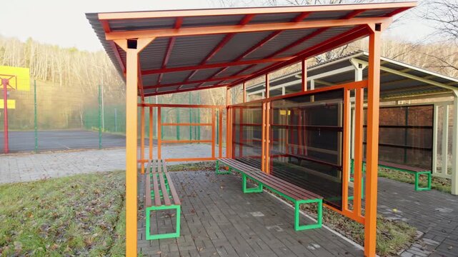 Vibrant shelters with benches line neatly paved pathway. Autumn leaves scatter across ground under tall bare trees and weather feels quiet
