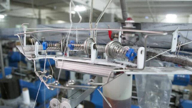 Thread passes through industrial yarn guide above automated knitting machine. Metal guides control steady thread movement during textile production.