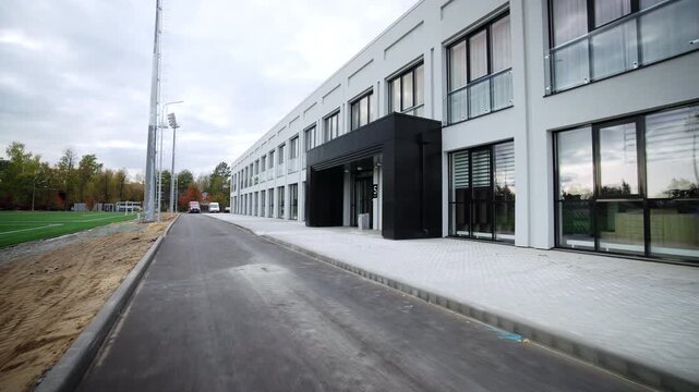 Asphalt driveway runs parallel to long modern facility facade. Smooth dark road surface contrasts with pale brick pavement path borders